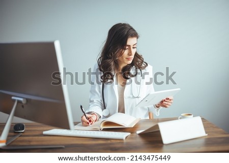 The mid adult female doctor reviews her patient's records on her computer in her office. Female doctor working at office desk, she is smiling, healthcare professionals