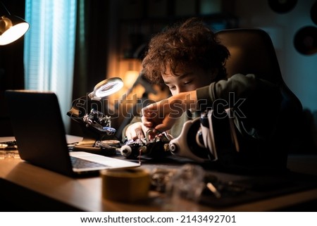 A young DIYer builds a robot. Child sits at desk and connects cables, toy electronics, solders wires, boy is interested in robotics Royalty-Free Stock Photo #2143492701