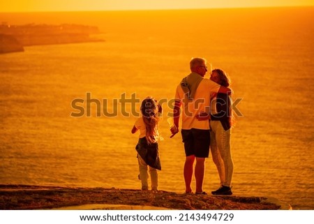 Silhouette of a young romantic couple with a child enjoying the evening on a cliff above the sea with a red burning sunset. Traveling family holding hands and standing on top of a mountain at sunset.