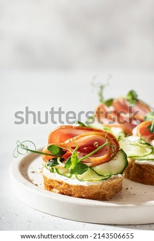 Close-up with Bruschetta with baguette, bacon or meat, cream cheese, micro-greenery, fresh cucumber and sprouts, on white plate on white textured background. Antipasto. Vertical orientation Royalty-Free Stock Photo #2143506655