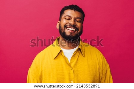 Happy young man smiling cheerfully with his eyes closed in a studio. Handsome young man with piercings standing alone against a pink background. Stylish young man feeling vibrant. Royalty-Free Stock Photo #2143532891