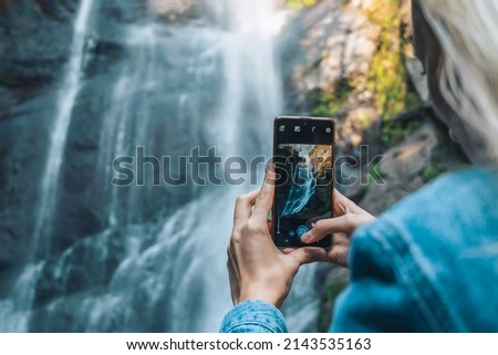 Close-up of a young female traveler photographing a waterfall with a mobile phone. First-person point of view, copy space. Concept of travel, tourism, blogging
