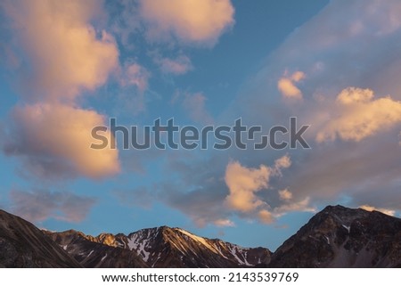 Scenic landscape with high snow mountain with sharp rocky pinnacle in golden sunlight under clouds of sunset color at changeable weather. Colorful view to large mountain top under orange clouds in sky