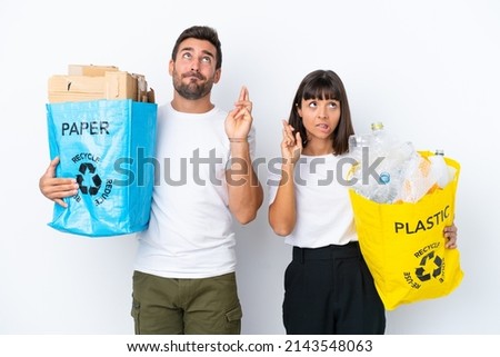 Young couple holding a bag full of plastic and paper to recycle isolated on white background with fingers crossing and wishing the best