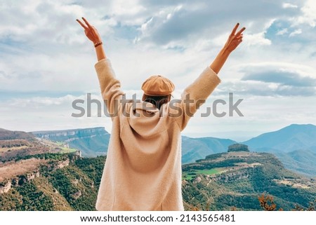 Back view young stylish hiker woman standing with raised hands enjoying on top rocky mountains, arms outstretched in victory sign gesture. Cloudy day.