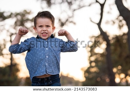 Funny pleased happy boy doing strength with his arms in the park outdoors in the sunset
