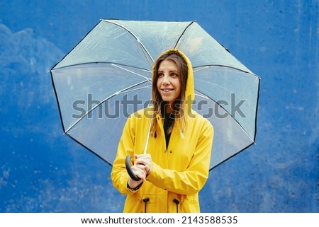 Mid waist portrait of caucasian woman in yellow waterproof clothes under rain with umbrella. Horizontal view of caucasian happy woman outdoors holding umbrella on blue background. Seasonal concept