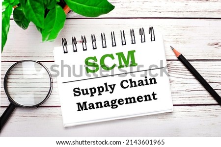 On a light wooden background, a magnifying glass, a pencil, a green plant and a white notebook with text SCM Supply Chain Management. Business concept