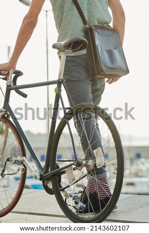 Crop of stylish man with leather crossbag walking with fixie bike along promenade. Royalty-Free Stock Photo #2143602107