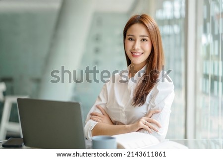 Confident young Asian business woman sitting with arms crossed smiling looking at camera in the office.
