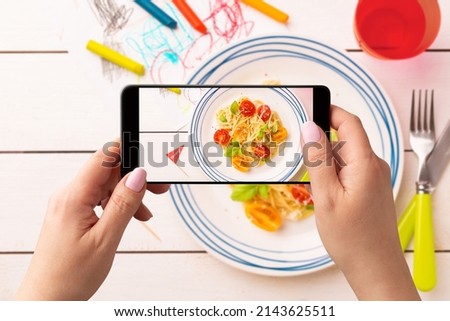 Woman taking photo of kid’s dinner (spaghetti) with smartphone. Food blogger using phone to capture meal. Lifestyle trend - posting and sharing food pictures on social media.