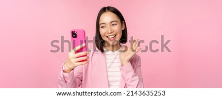 Stylish asian businesswoman, girl in suit taking selfie on smartphone, video chat with mobile phone app, posing against pink studio background