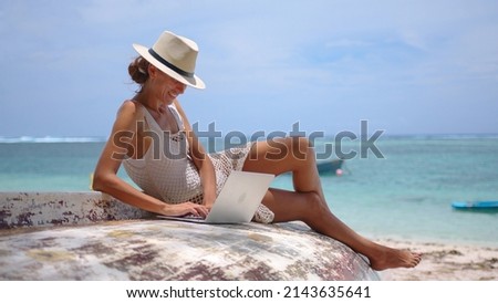 Business woman in yellow hat using laptop computer on the background of a tropical beach with turquoise water. Vacation, freelance job, leisure activity concept