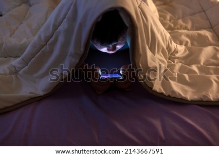 Little boy on cell phone under blanket. Curious boy sits in bed under white blanket and play games on smartphone in the dark room.  Image of nice boy in pajama sitting in front of mobile. 