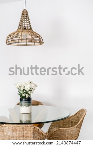 Vertical view of wicker armchair with bamboo material near glass table in kitchen. Ceiling light lamp and chair with rattan in cozy apartment with boho style room. Home decor, fresh flowers in vase