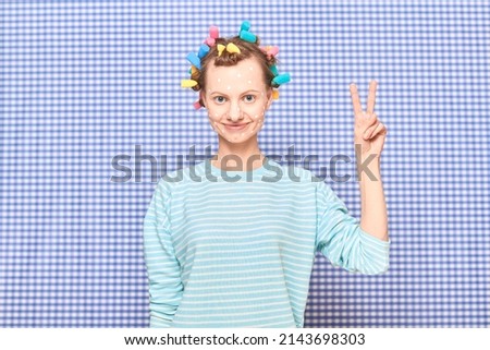 Portrait of happy young woman with white spots of skincare product on face, with colorful hair curlers on head, showing peace or victory sign, standing over shower curtain background in bathroom