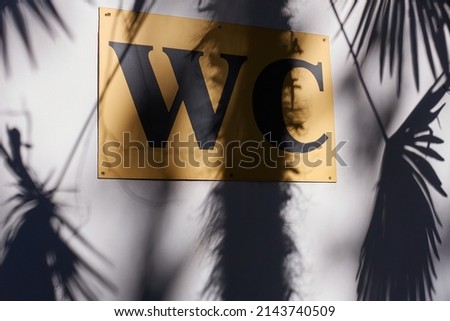 The shadow of a palm tree falls on a white wall with a yellow sign that says water closet in black letters.