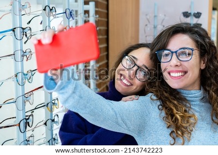 Two happy female friends in a optic shop wearing a pair of glasses while taking a photo with their smartphone Royalty-Free Stock Photo #2143742223