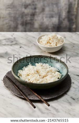 Still life photography of stoneware bowls, underplate and chopsticks with steamed white rice on a mottled concrete background for food photography. 