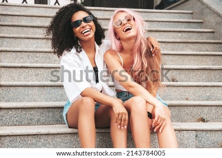 Two young beautiful smiling hipster female in trendy summer clothes.Sexy carefree multiracial women posing on the street background.Positive models having fun in sunglasses. Sitting at the stairs Royalty-Free Stock Photo #2143784005