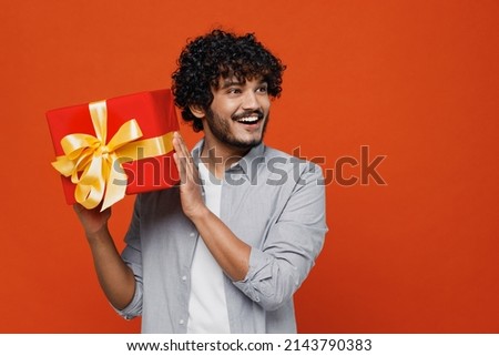 Cheerful happy smiling blithesome happy young bearded Indian man 20s years old wears blue shirt hold raise up red present box with gift ribbon bow isolated on plain orange background studio portrait