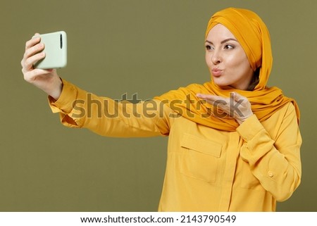 Young girlfriend arabian asian muslim woman 20s wear abaya hijab yellow clothes do selfie shot on mobile phone blowing air kiss isolated on olive green background People uae islam religious concept.