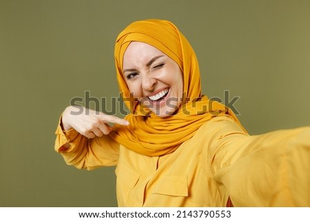 Close up fun young arabian asian muslim woman in abaya hijab yellow clothes do selfie shot on mobile phone point finger on herself isolated on olive green background People uae islam religious concept