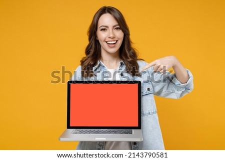 Young smiling happy satisfied fun woman 20s in denim shirt white t-shirt point index finger on laptop pc computer with blank screen workspace area chat isolated on yellow background studio portrait.