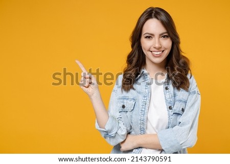Young smiling woman promoter in denim shirt white t-shirt recommend suggest select advert point index finger aside on workspace commercial promo area mock up copy space isolated on yellow background Royalty-Free Stock Photo #2143790591