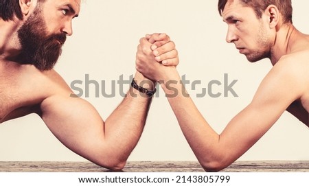 Heavily muscled bearded man arm wrestling a puny weak man. Arms wrestling thin hand, big strong arm in studio.