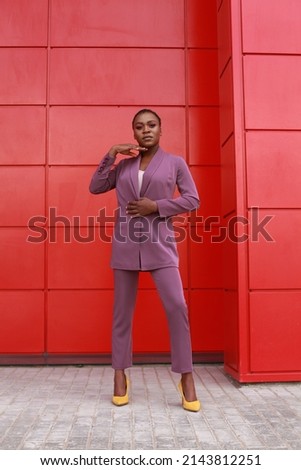 Black young female model posing in purple pants suit on urban red wall background. Looking formal but with a touch of elegance. Street style fashion image with copy space. Royalty-Free Stock Photo #2143812251
