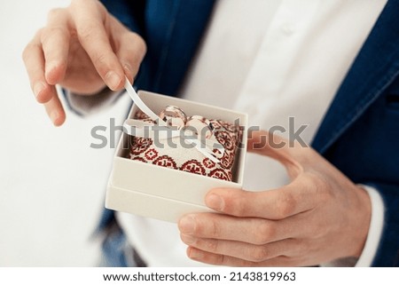 The groom holds gold wedding rings in a decorative box with a small pillow and ribbons for the bride in the morning indoors on a white background on the wedding day