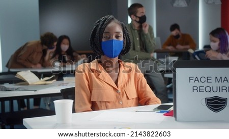 A young african professor is sitting in a blue protective mask with her students in the classroom and looking at the camera