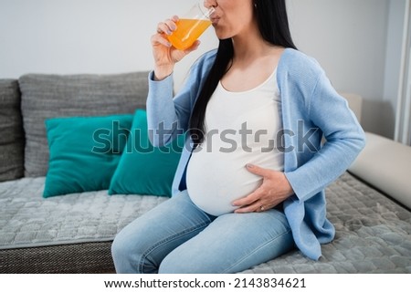 A pregnant woman drinks orange juice in the living room and holds her huge belly with her left hand. She sits on the sofa and enjoys drinking the necessary vitamins.Green pillows are in the background