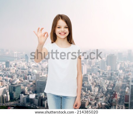 advertising, childhood, gesture and people concept - smiling little girl in white blank t-shirt showing ok sign over city background