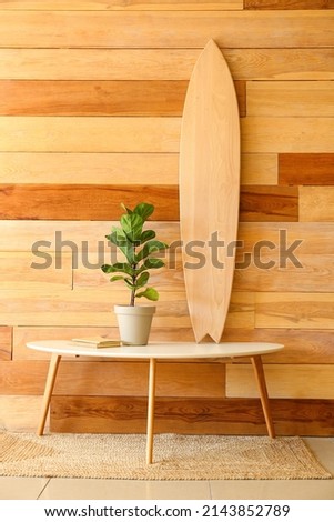 Table with surfboard, houseplant and books near wooden wall