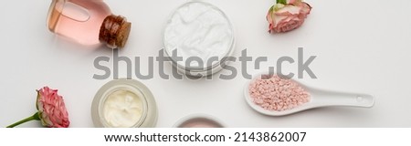 top view of flowers near pink sea salt, bottle with rose water and containers with cream on white, banner