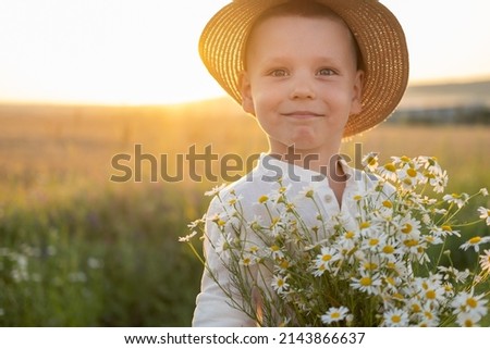 cute smiling child boy in straw hat holds bouquet of field chamomiles on sunset