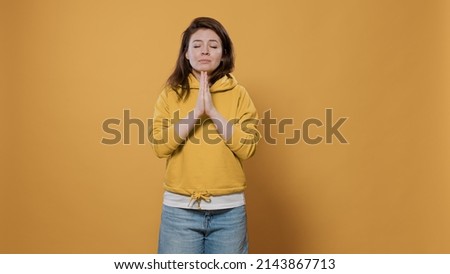 Humble young person wearing hoodie being religious having hope and faith with palms close together in studio. Casual woman feeling worried, looking up praying and asking for forgiveness.