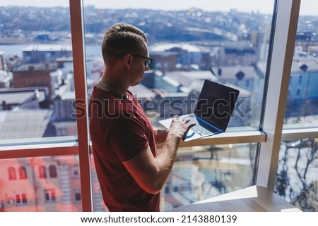 Smiling businessman stands by the window while working on a laptop. Multitasking concept. A busy freelancer in glasses holds a laptop in his hands while standing near the window. Smart casual.