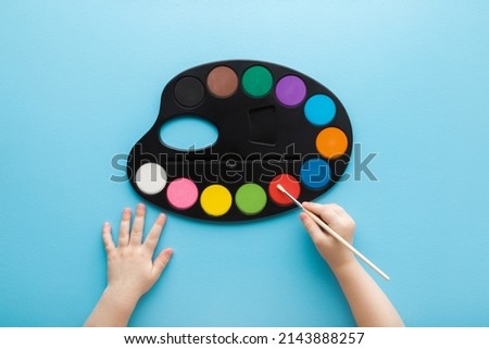 Baby hand holding paint brush and using colorful palette on light blue table background. Pastel color. Closeup. Point of view shot. 