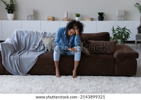 Depressed exhausted young African woman feeling stress, suffering from headache, sitting on cough at home, touching head, going through apathy, grief, loss. Depression, fatigue concept Royalty-Free Stock Photo #2143899321