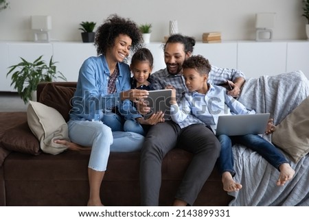 Cheerful young Black parents and curious little gen Z boy and girl resting together on home sofa, using laptop, tablet computer gadget, shopping internet, chatting online on social media Royalty-Free Stock Photo #2143899331
