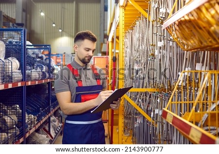 Male worker, salesman or building materials store manager conducts inventory of goods. Young man in overalls writes on clipboard available stock of goods in hardware or construction store. Royalty-Free Stock Photo #2143900777