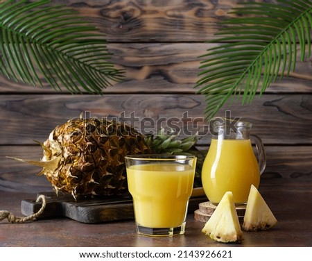 A clear glass of pineapple juice on a brown table. A whole pineapple on a wooden plank in the background. A jug of juice on the table. Palm leaves in the background