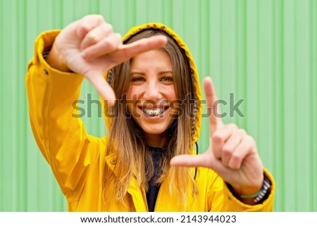 Front cropped view of woman with yellow raincoat doing a snap photography gesture. Horizontal view of woman framing her head with fingers in green wall. People isolated in background with copy space.
