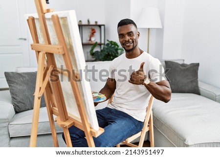 Young african man painting on canvas at home doing happy thumbs up gesture with hand. approving expression looking at the camera showing success. 
