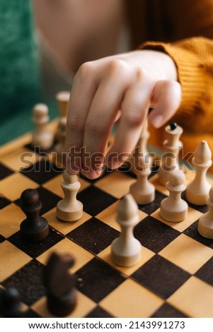 Vertical shot of unrecognizable woman making chess move with piece sitting at table in dark room, selective focus. Pretty intelligent lady playing logical board game alone at home, blurred background.