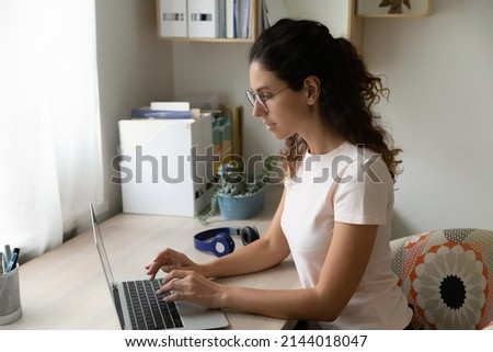 Busy young Hispanic business woman sit at desk wear glasses work on laptop texting e-mail solve business from homeoffice looking serious and confident, make telecommuting remote job from home concept Royalty-Free Stock Photo #2144018047