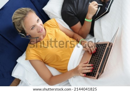 Woman operator in headphones works on laptop at home on bed. Remote work and telework concept